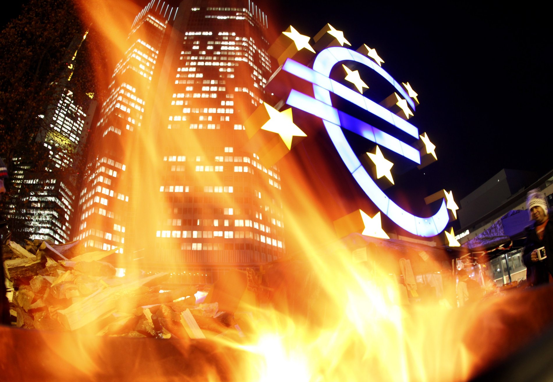 Znak eura u sídla ECB ozářený ohněm při protestech ve Frankfurtu
